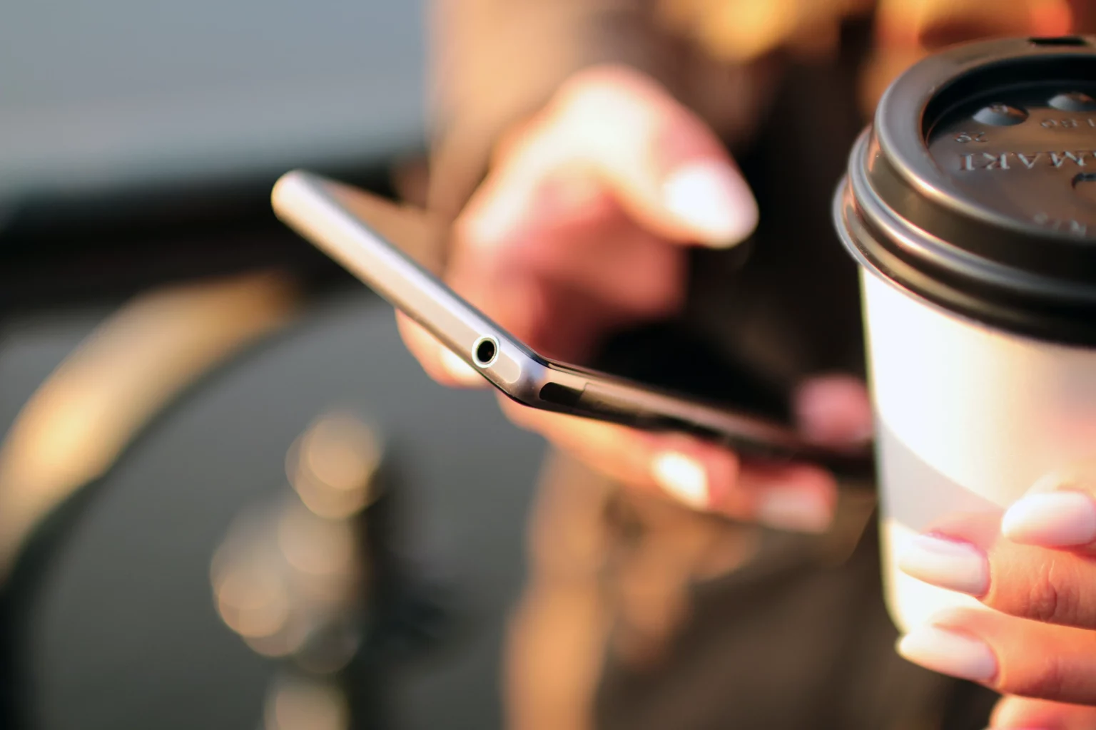 A close-up of a person's hands holding a smartphone and a takeaway coffee cup against a blurred bright background somewhere outdoors.