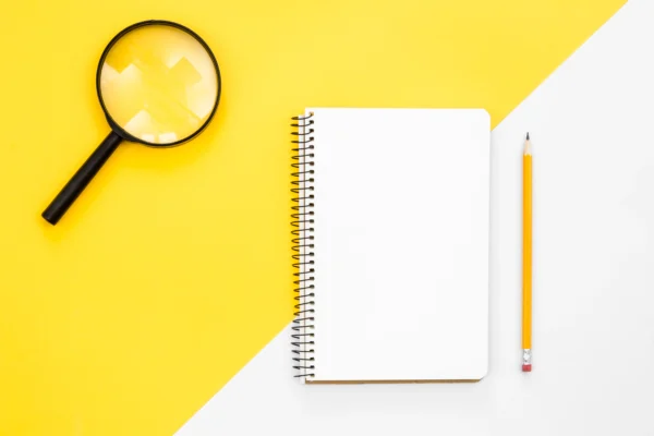 A magnifying glass laying on a vibrant yellow surface next to an open notebook and a pencil.